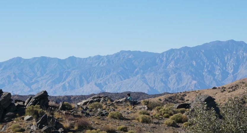 A person sits in solitude in a desert landscape. There are mountains in the background. 